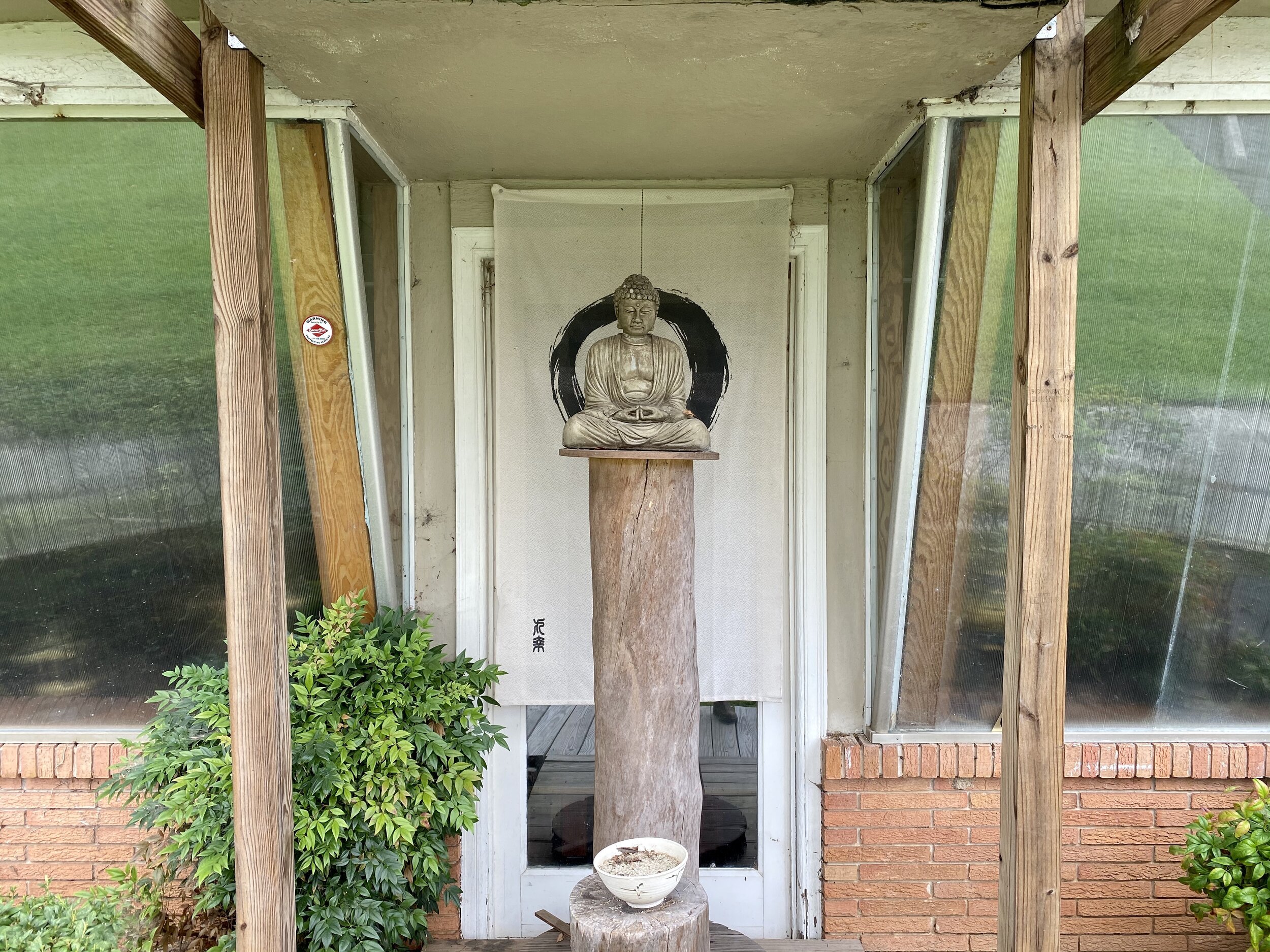 A statue of the Buddha seated in meditation atop a tall pole in front of a hanging of an enso outside of a rustic Zen center