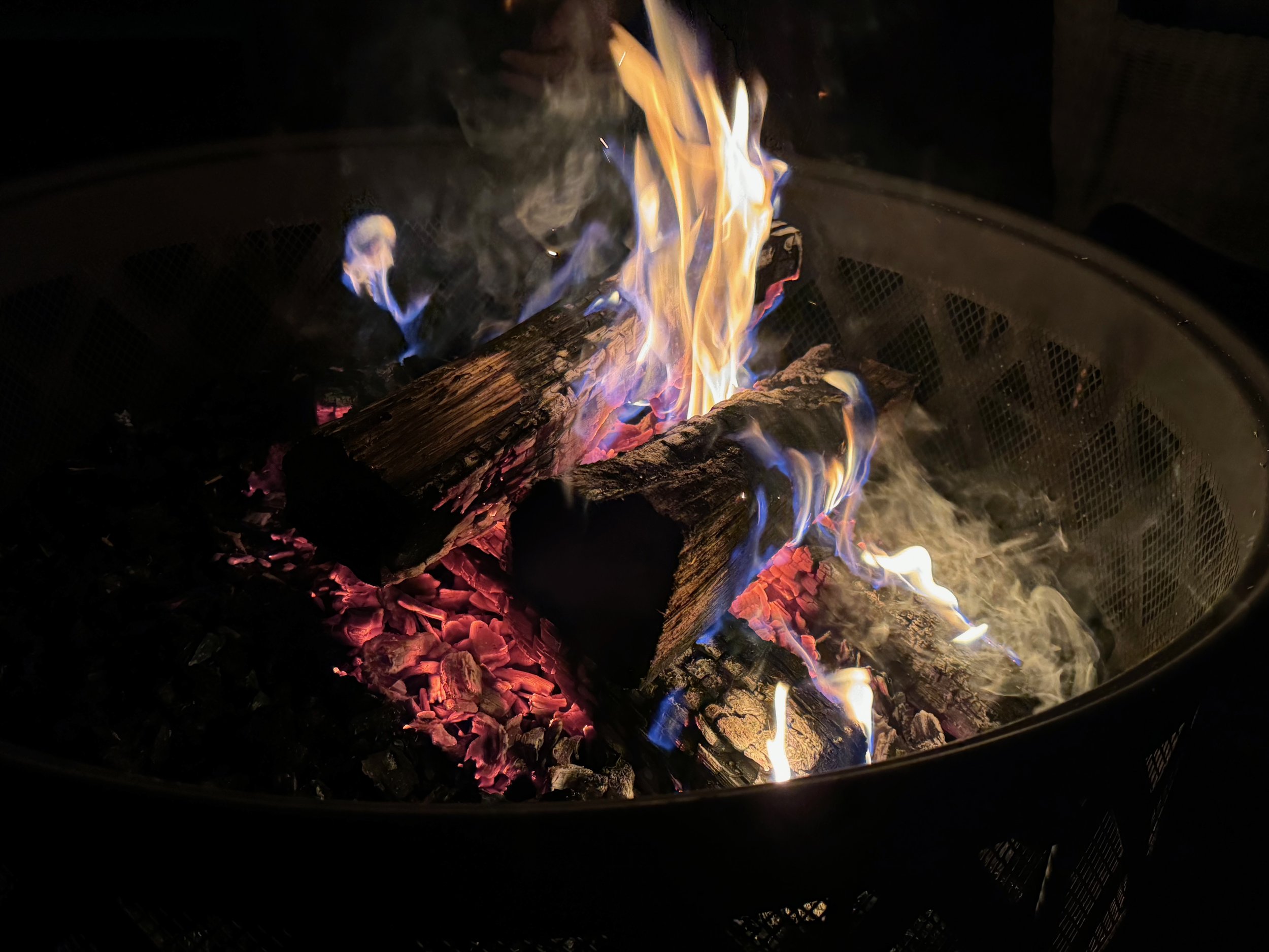 A small fire in an outdoor fire pit with four logs laid crosswise on a bed of coals